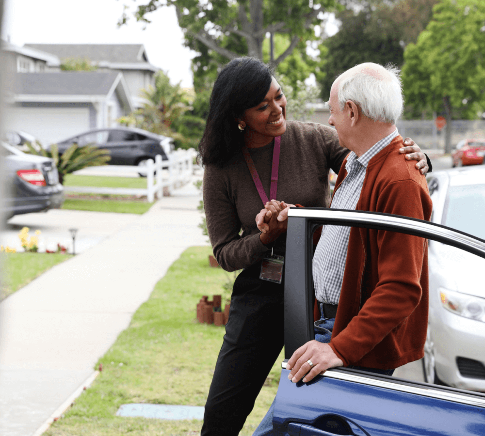 A woman helps an elderly man out of a car in a suburban neighborhood, both smiling and holding hands. - Home Instead