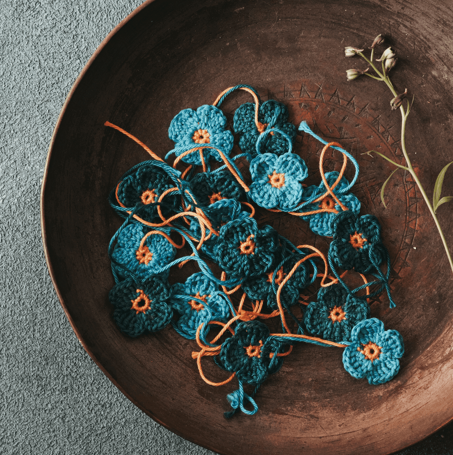 Crocheted blue flowers with orange centers in a brown dish, beside green stems on a textured gray surface. - Home Instead