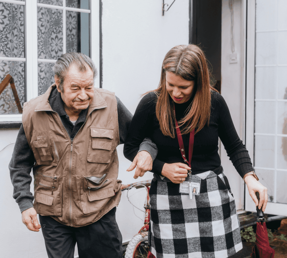 A caregiver assists an elderly man outside a house, holding his arm while he holds a cane. - Home Instead