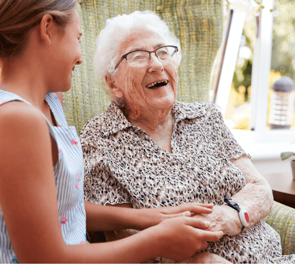 A smiling older woman wearing glasses sits and laughs with a girl in a patterned dress who is holding her hand. - Home Instead
