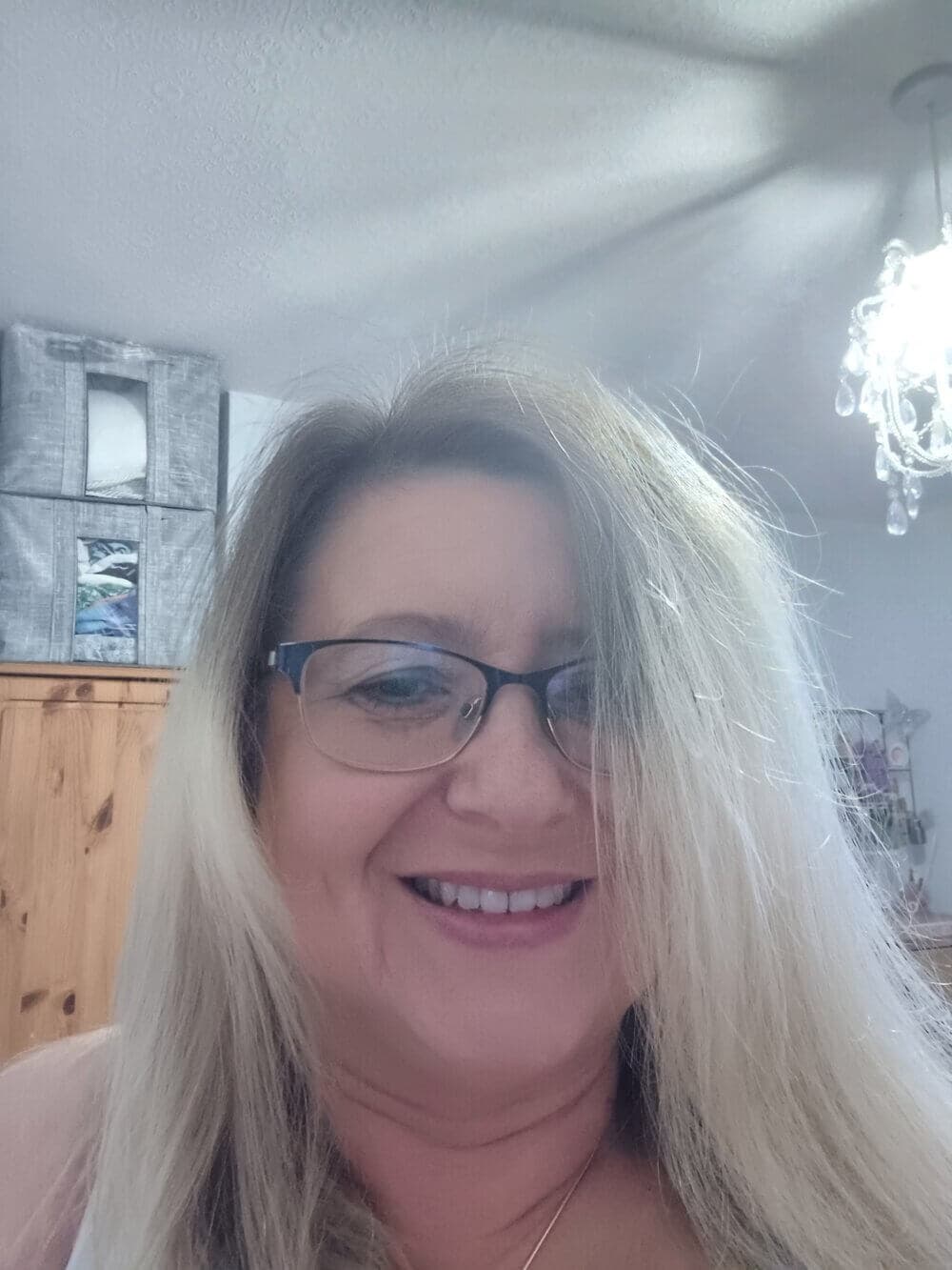 A woman with glasses and long blonde hair smiling indoors, with a chandelier and wooden cabinet in the background. - Home Instead