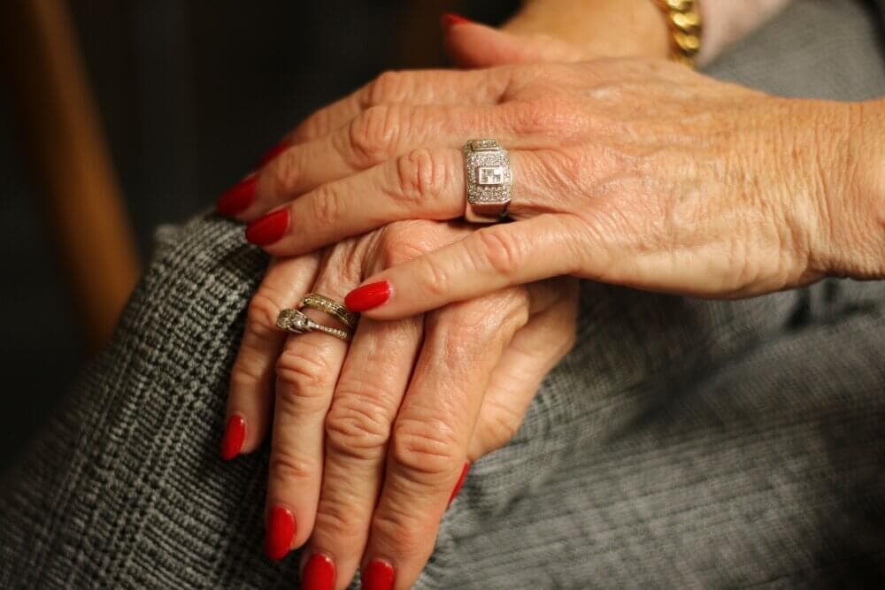 Elderly hands with red nails and two diamond rings resting on a grey patterned surface. - Home Instead