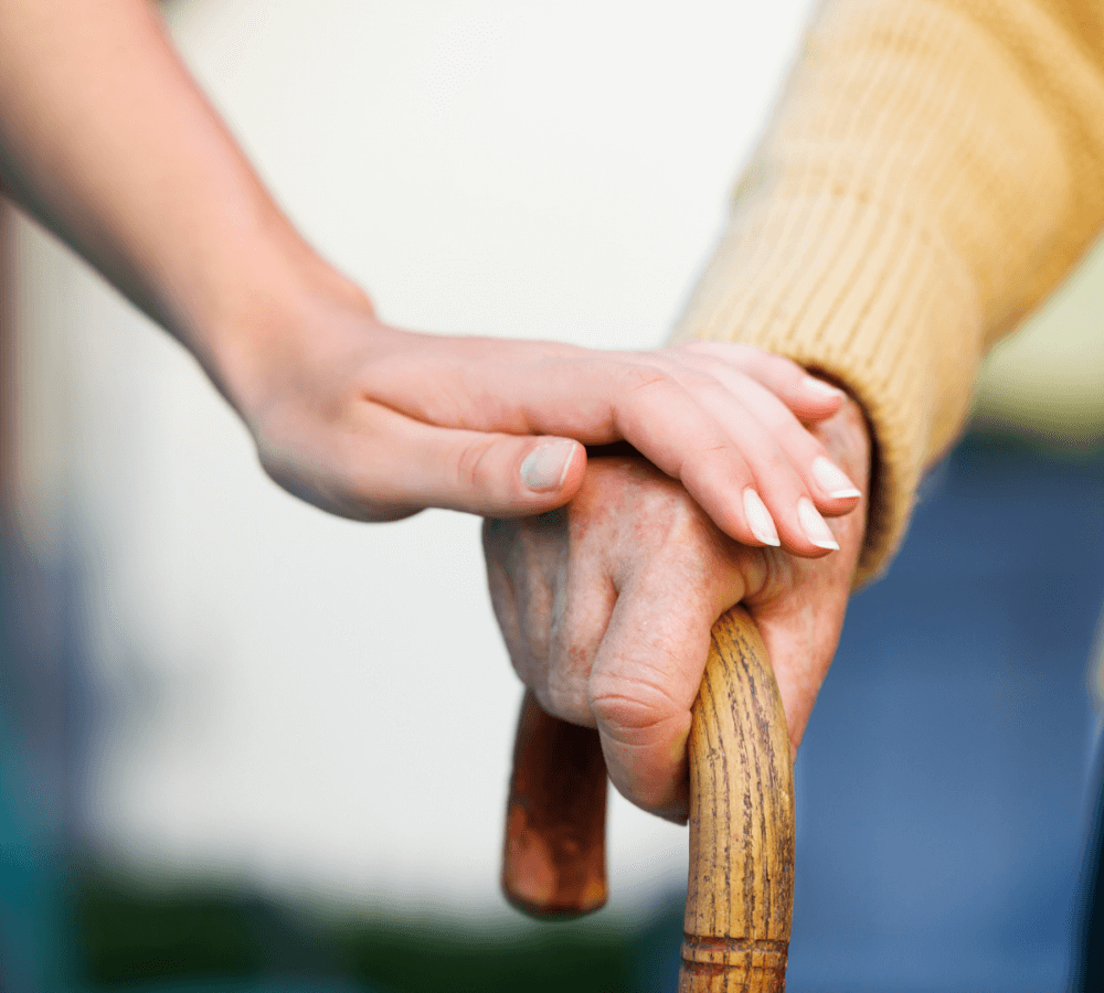 A younger hand rests on an elderly hand holding a wooden cane, symbolizing support and care. - Home Instead
