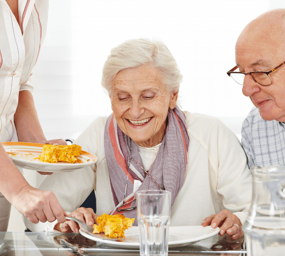 Three elderly individuals sharing a meal at a well-lit dining table, with one being served a piece of casserole. - Home Instead