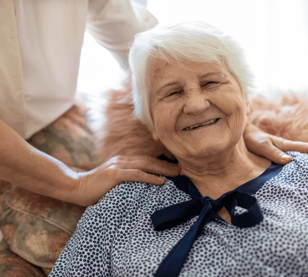 An elderly woman with white hair smiles warmly while a person's hand rests gently on her shoulder. - Home Instead