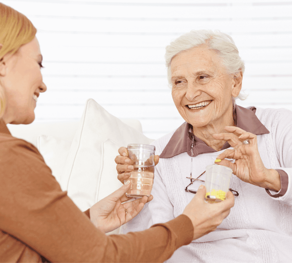 A young woman hands water and medication to an elderly smiling woman sitting on a couch. - Home Instead