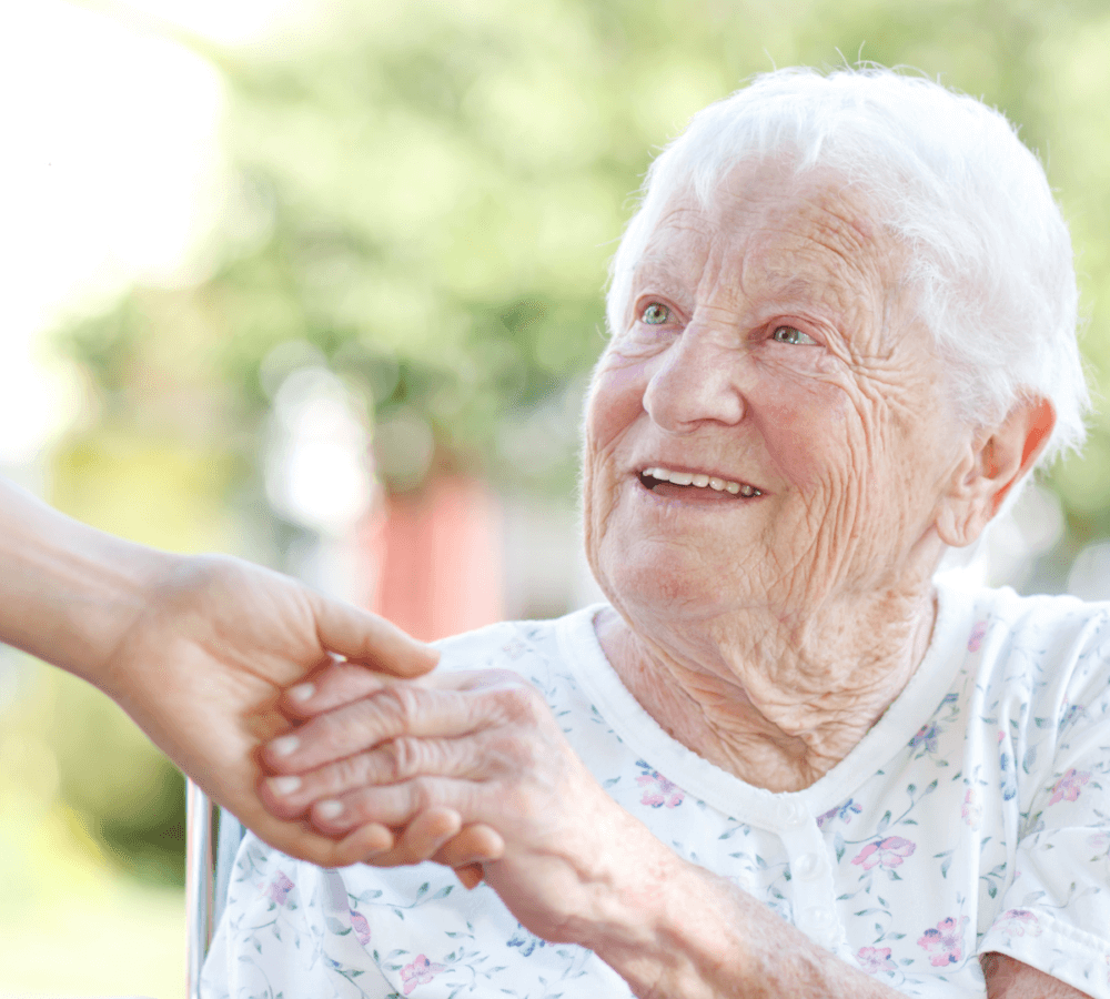 Elderly woman with white hair smiling and holding hands with another person outdoors. - Home Instead