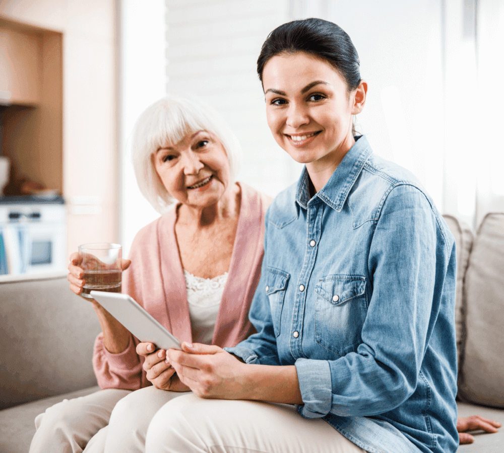 Young woman with tablet sits beside smiling elderly woman holding a glass of water on a couch in a cozy room. - Home Instead