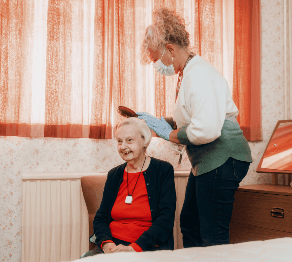A caregiver in a mask and gloves brushes an elderly woman's hair in a cozy room with orange curtains. - Home Instead