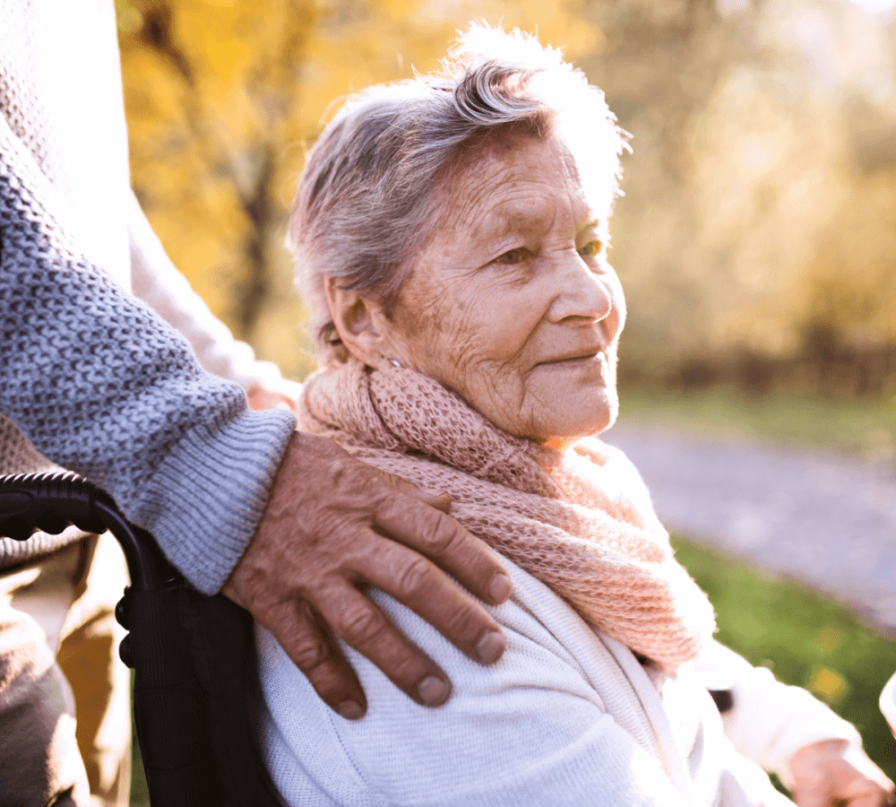Elderly woman in a wheelchair outdoors, with a person's hand gently resting on her shoulder, surrounded by autumn trees. - Home Instead