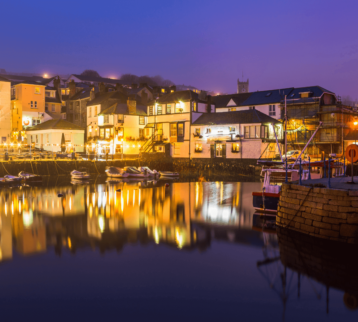 A serene coastal village at dusk with illuminated boats, houses, and buildings reflecting on calm waters. - Home Instead