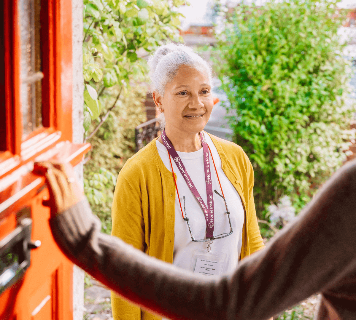 A woman wearing a yellow cardigan and name badge stands at a red door, smiling at someone opening it. - Home Instead