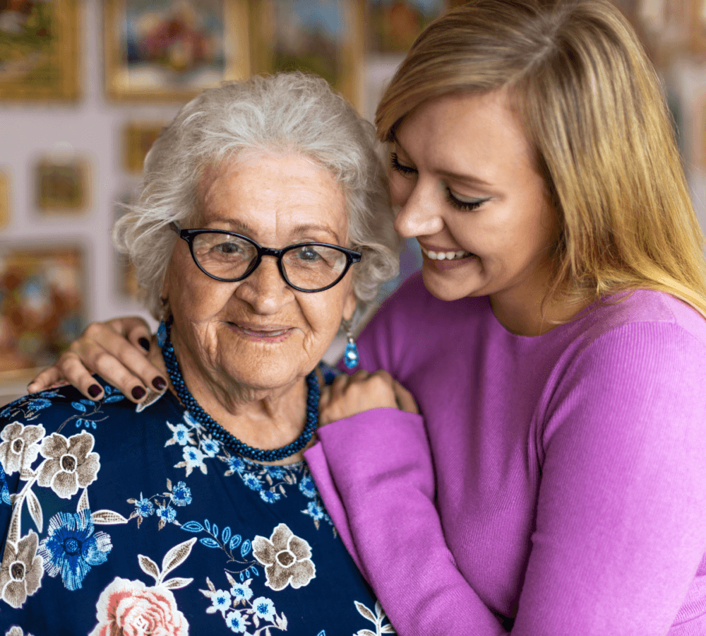 An elderly woman and a younger woman smile as they embrace, the younger woman wearing a purple sweater. - Home Instead