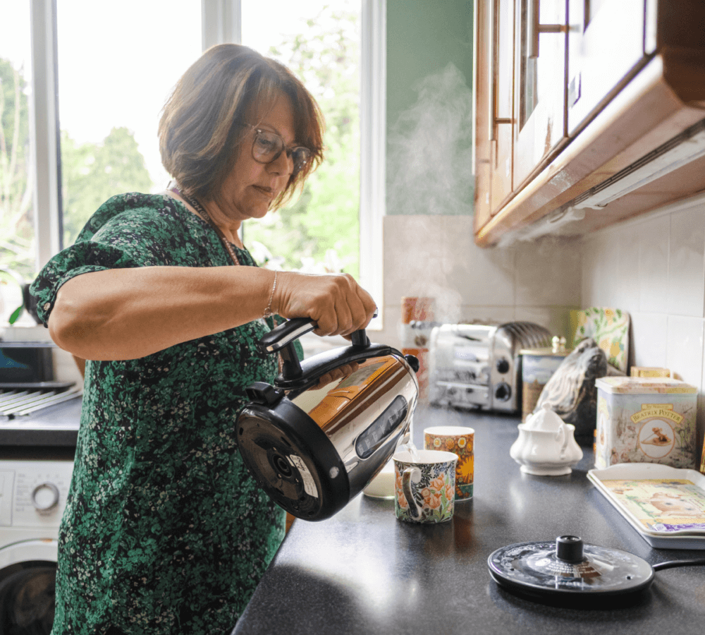 A woman pours hot water from an electric kettle into a teacup in a kitchen with various items on the counter. - Home Instead
