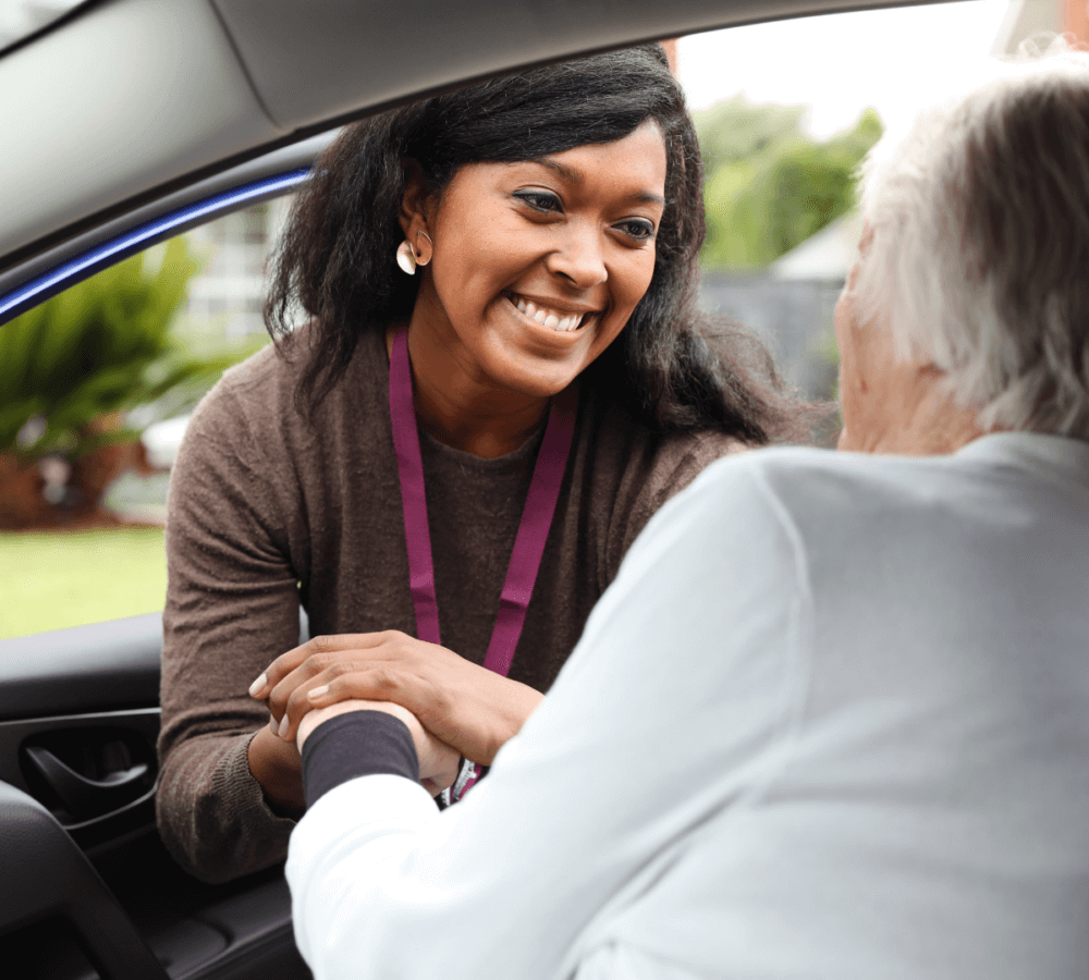 A smiling woman greeting and shaking hands with an elderly person sitting in a car. - Home Instead