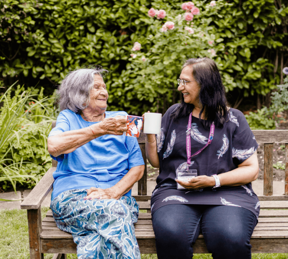Two women sitting on a bench in a garden, smiling and clinking mugs together. - Home Instead