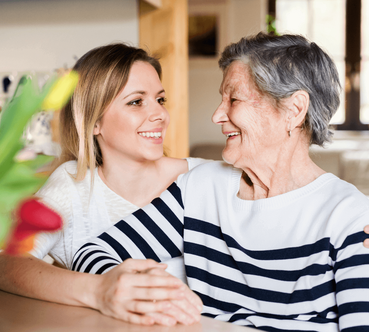 Young woman and elderly woman smile and embrace at a table with a bouquet of flowers in the foreground. - Home Instead
