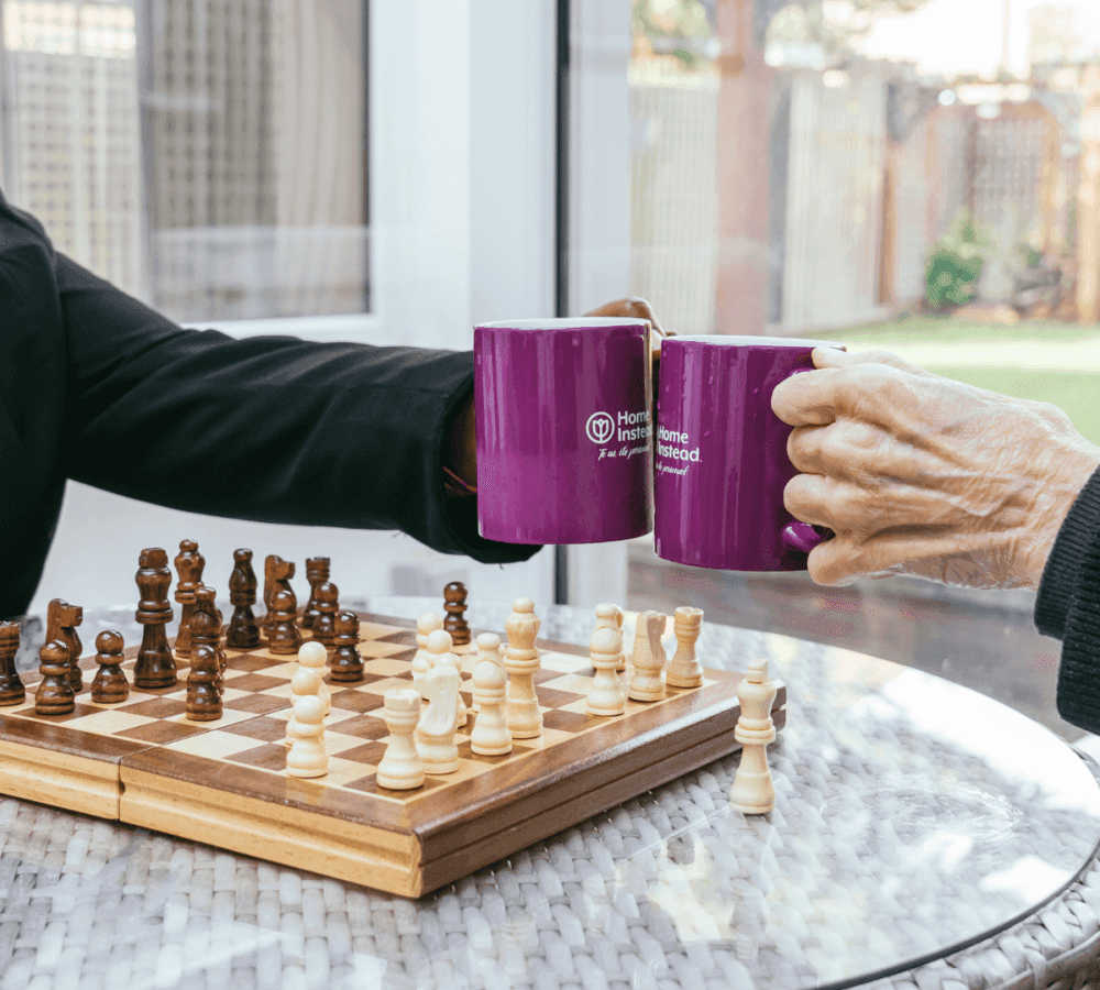 Two people clink purple mugs over a chessboard with pieces mid-game, seated at a glass-top table. - Home Instead