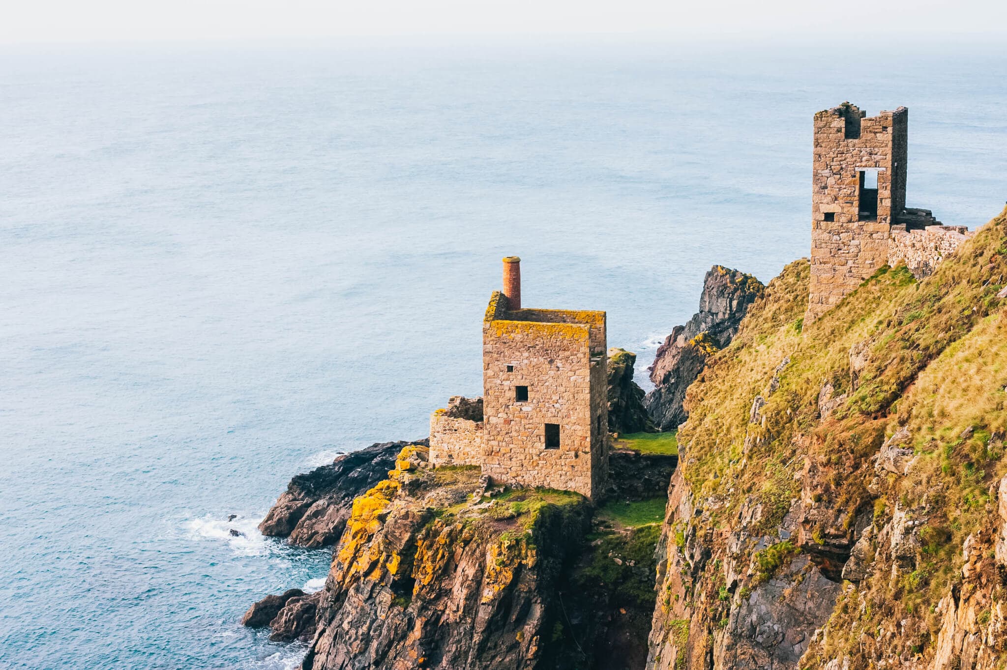Two stone buildings on a grassy cliff overlooking the ocean, with waves breaking on the rocks below. - Home Instead