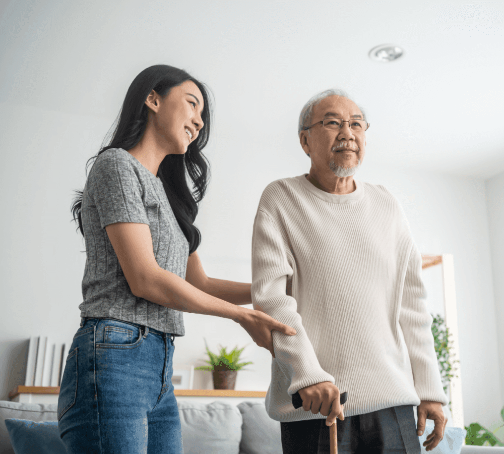 Young woman assisting an elderly man with a cane inside a cozy, light-filled living room. - Home Instead
