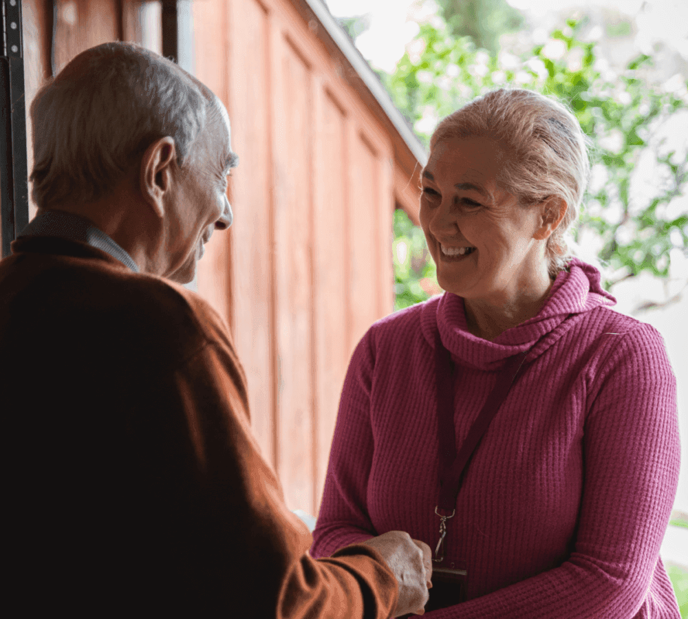 Two elderly people smiling and shaking hands indoors, with a wooden wall and greenery in the background. - Home Instead