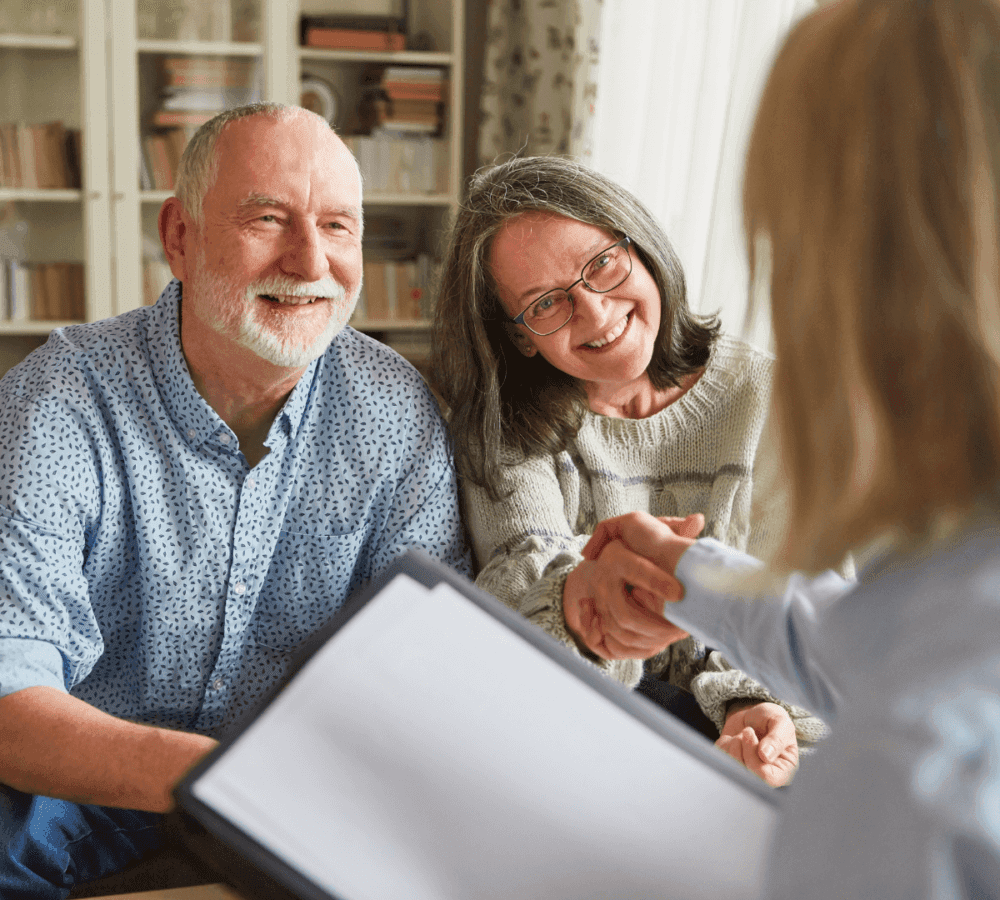 Smiling older couple sitting on a couch, shaking hands with a person holding a document in the foreground. - Home Instead