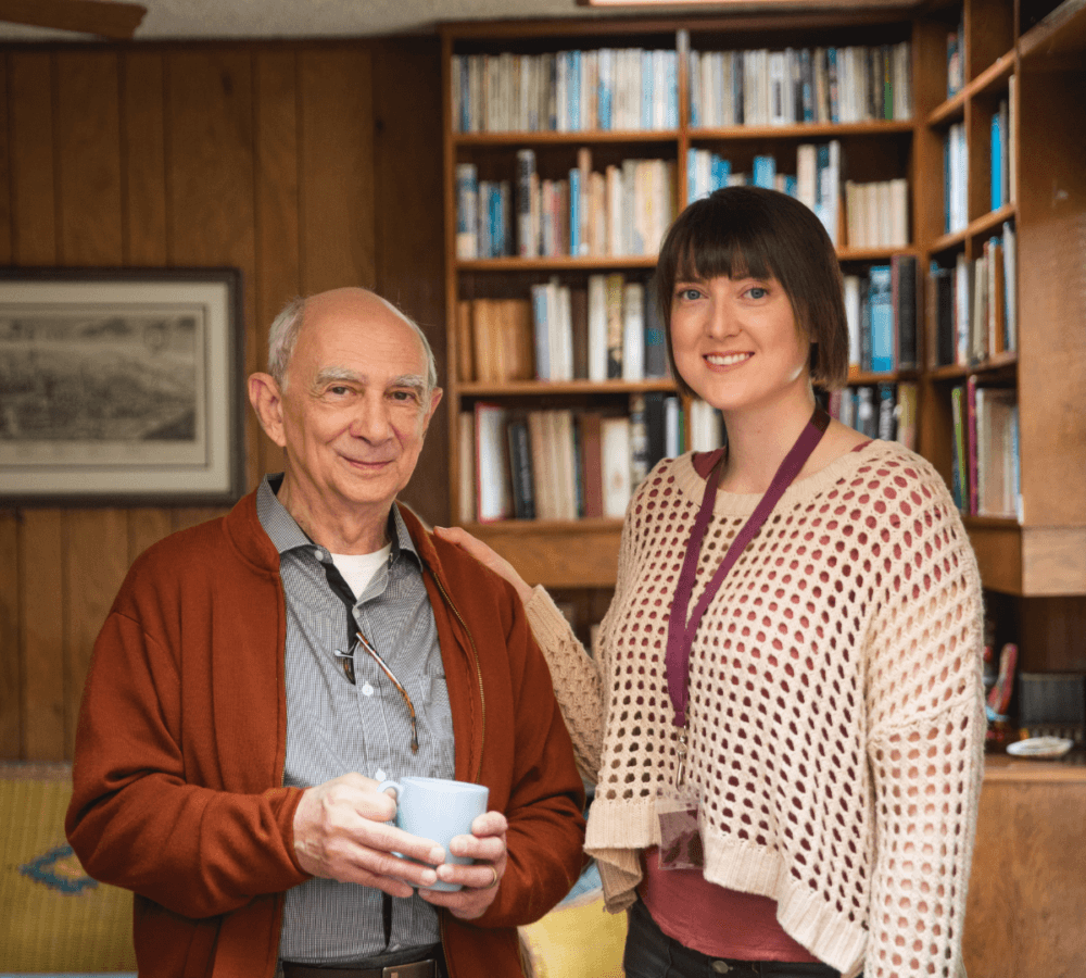 An elderly man holding a mug and a smiling young woman stand in front of a bookshelf in a cozy room. - Home Instead
