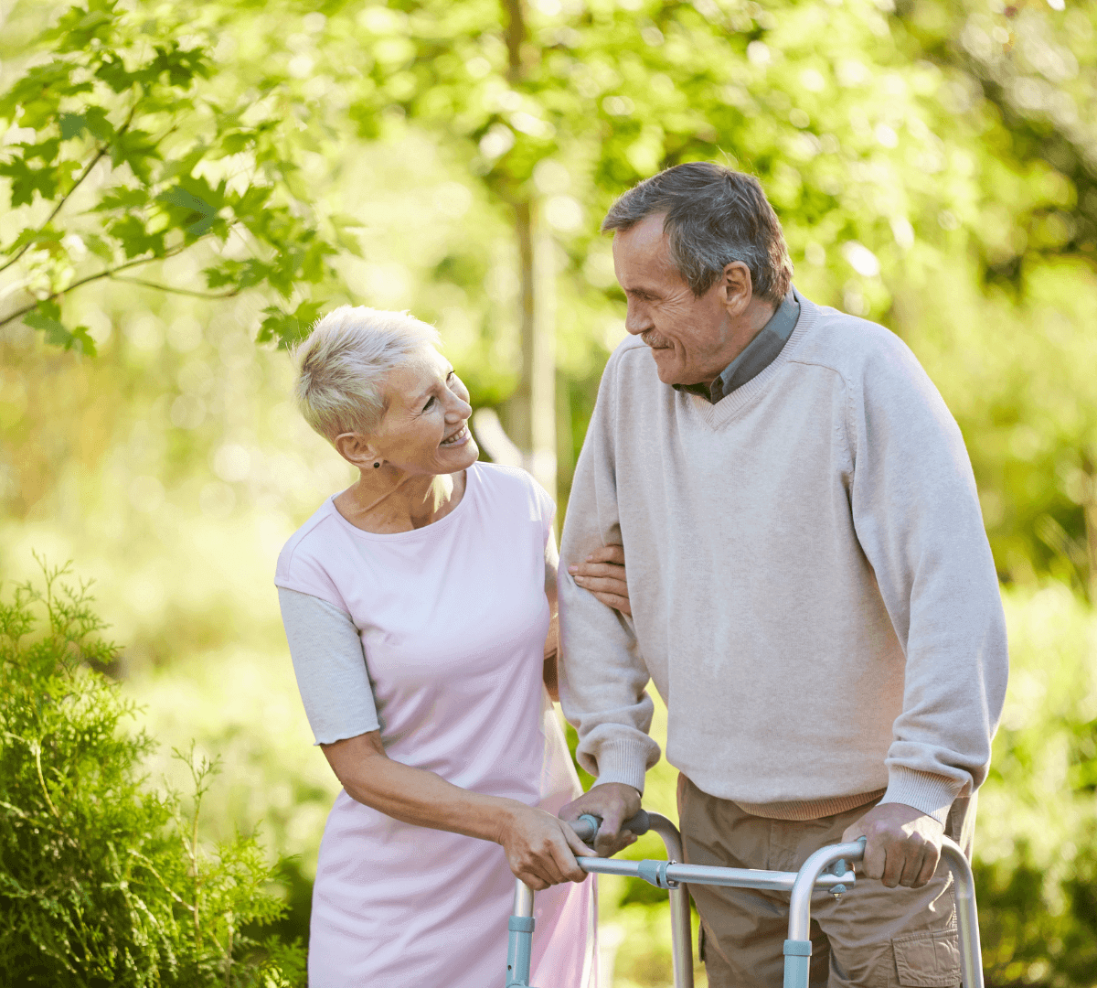 An elderly couple smiles and walks together, the woman supporting the man as he uses a walker in a garden. - Home Instead