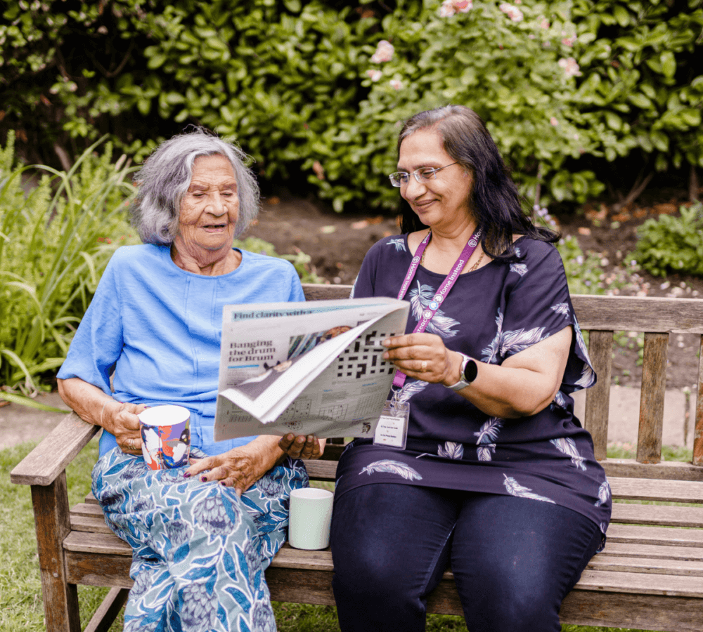 Two women sitting on a bench in a garden, one reading a newspaper and the other holding a mug, sharing a laugh. - Home Instead