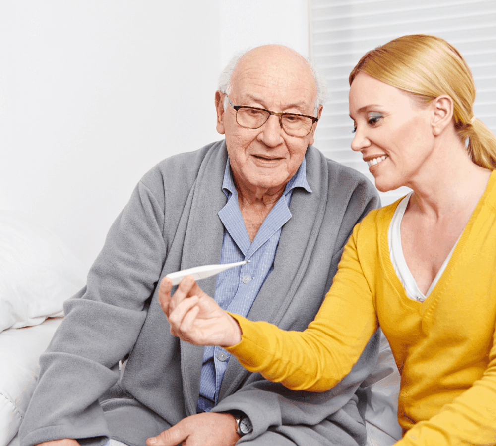 Smiling woman showing a thermometer to an elderly man in a gray robe, both seated on a bed. - Home Instead