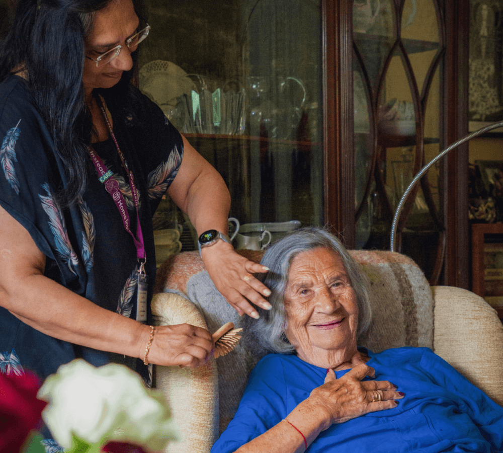 A caregiver combs the hair of a smiling elderly woman sitting in a comfy chair, both appearing content. - Home Instead