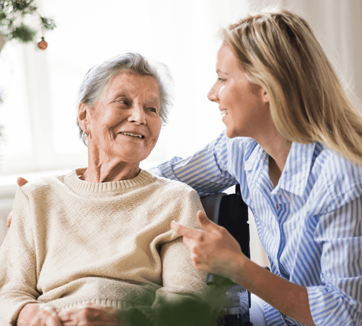 Young woman smiling and talking with an older woman in a sweater, both looking happy in a well-lit room. - Home Instead