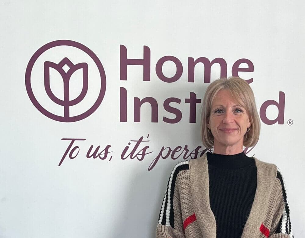 A smiling woman stands in front of a wall with the Home Instead logo and the tagline "To us, it's personal. - Home Instead