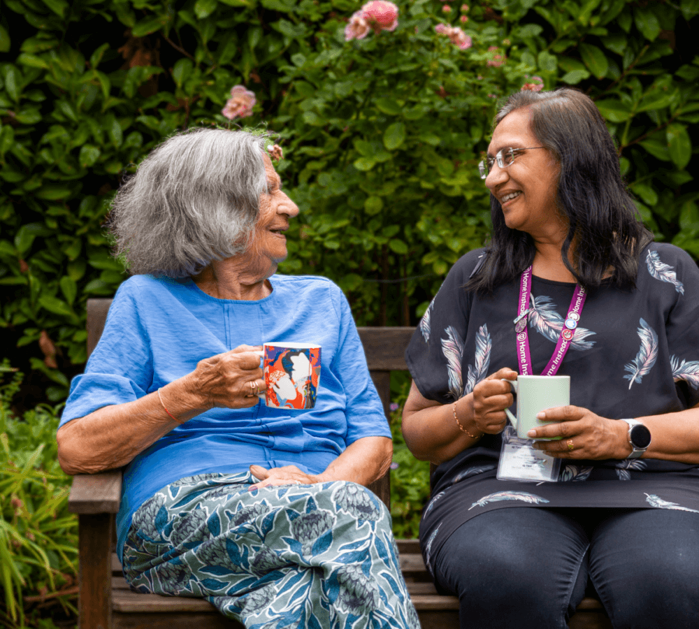 Two women sitting on a bench, smiling and holding mugs, with greenery and flowers in the background. - Home Instead