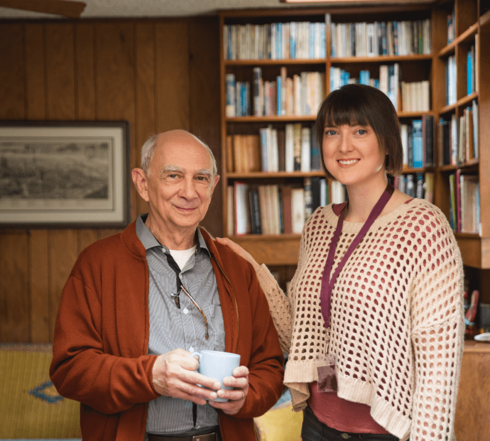 An older man holding a mug and a younger woman with a lanyard stand smiling in a wooden-paneled room with bookshelves. - Home Instead
