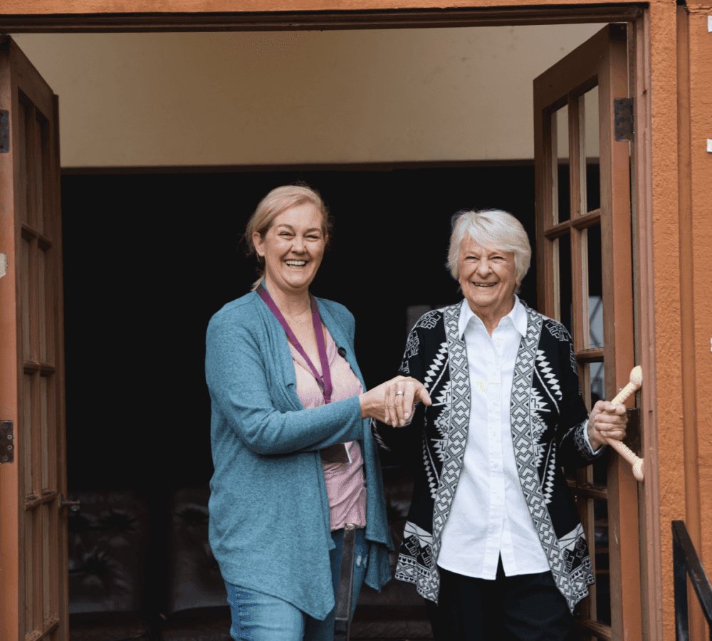 Two women smiling and holding hands while standing in a doorway; one holds a cane. - Home Instead
