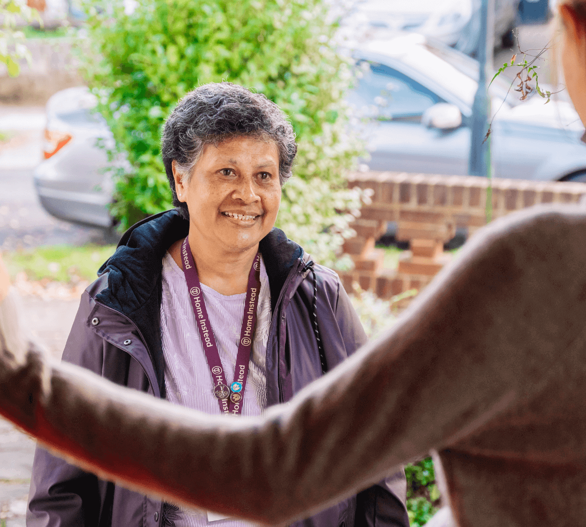 A person smiles while greeting someone at an open door, wearing a jacket and lanyard, with greenery in the background. - Home Instead