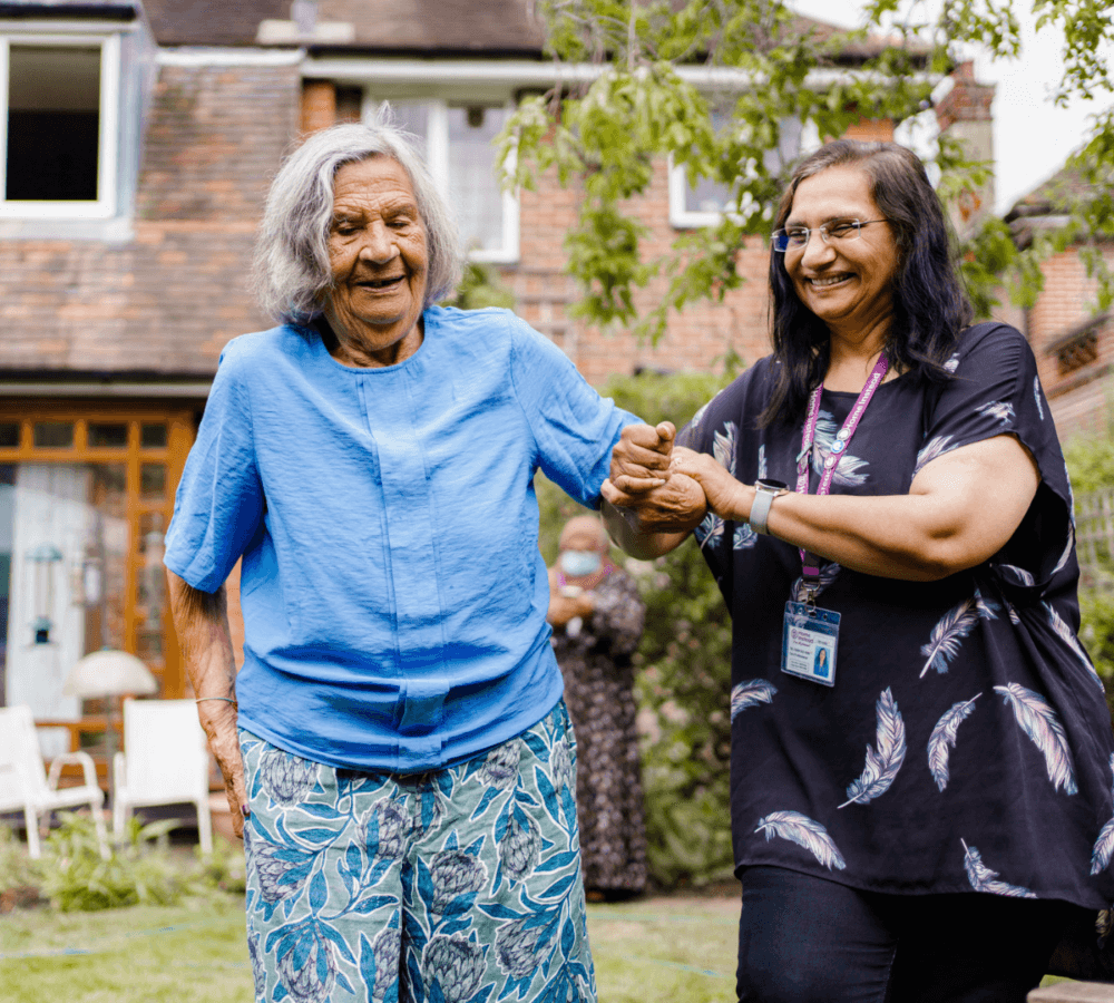 Two women, one elderly, walking in a garden while smiling and holding hands in a joyful moment. - Home Instead