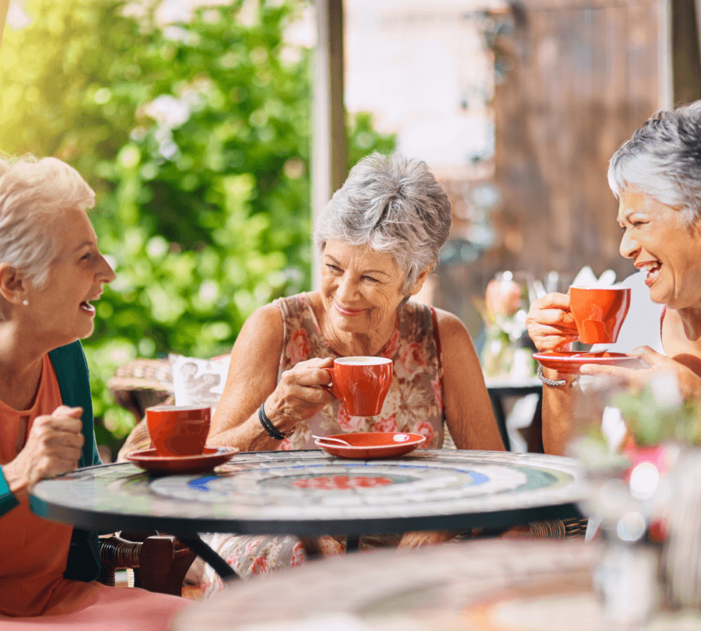 Three elderly women enjoying tea and laughing together at an outdoor café, holding red cups and sitting at a round table. - Home Instead