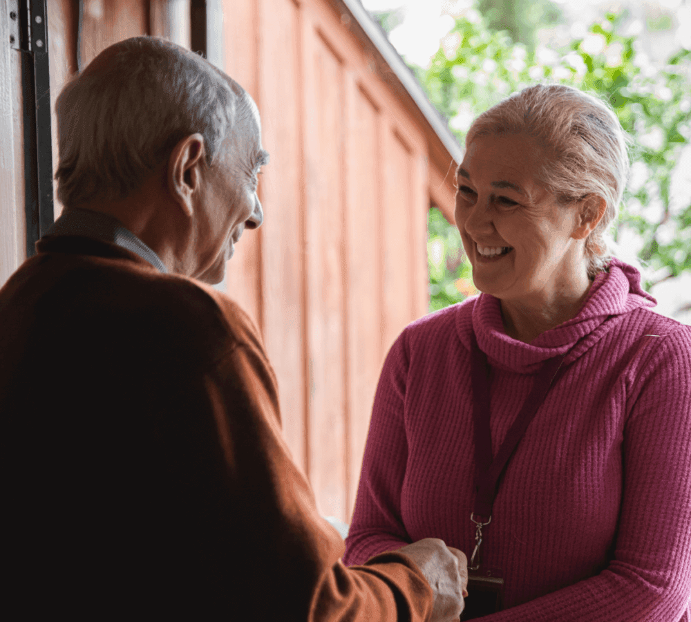 Two smiling older adults, a man and a woman, shaking hands outside near a wooden building. - Home Instead