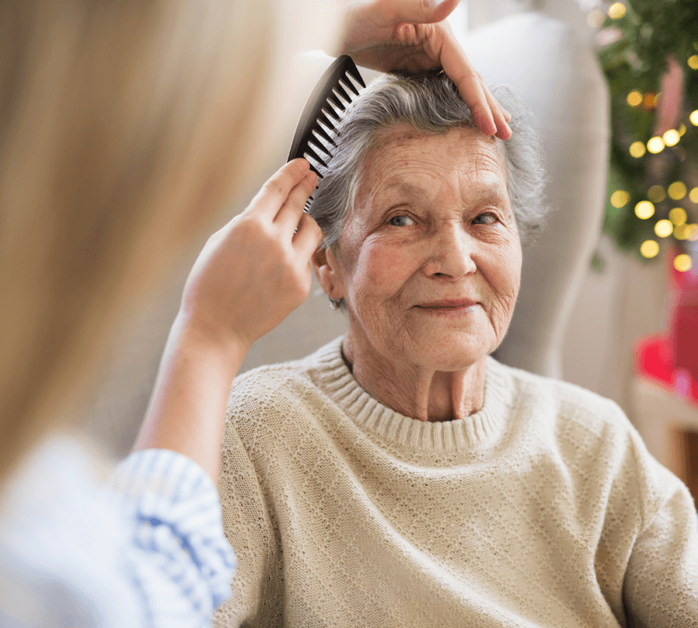 An elderly woman smiles as someone combs her hair, with holiday decorations in the background. - Home Instead