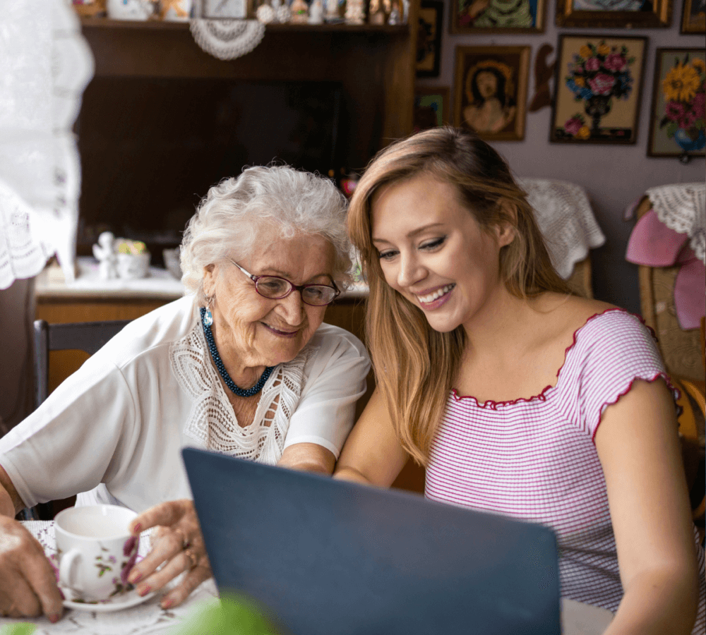 A smiling elderly woman and a younger woman sit together at a table, looking at a laptop screen. - Home Instead