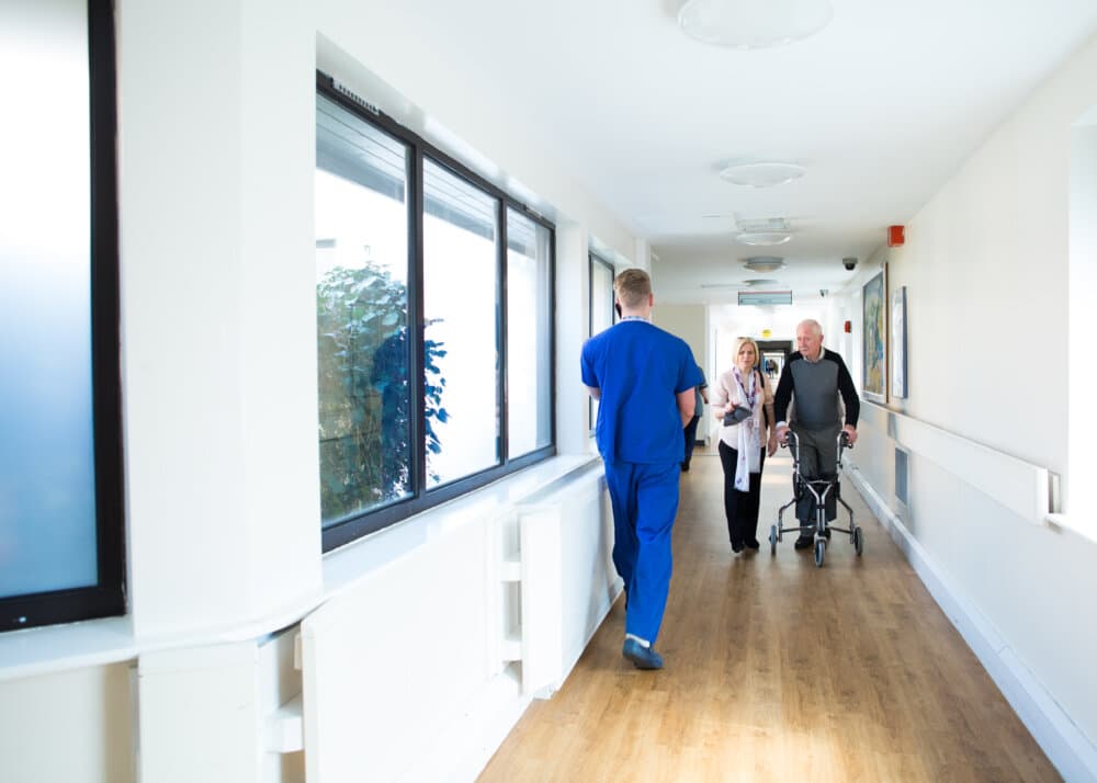 A healthcare worker in blue scrubs walks down a hospital hallway towards an elderly man using a walker and a woman. - Home Instead