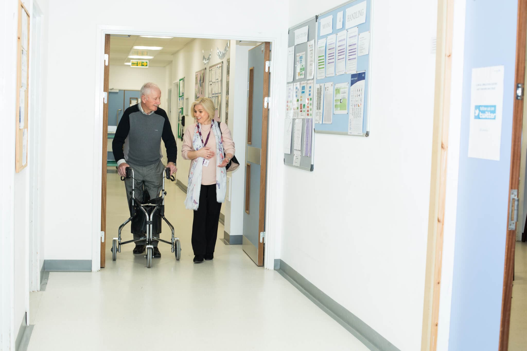 An elderly man using a walker is accompanied by a woman in a hospital hallway. They appear to be having a conversation. - Home Instead