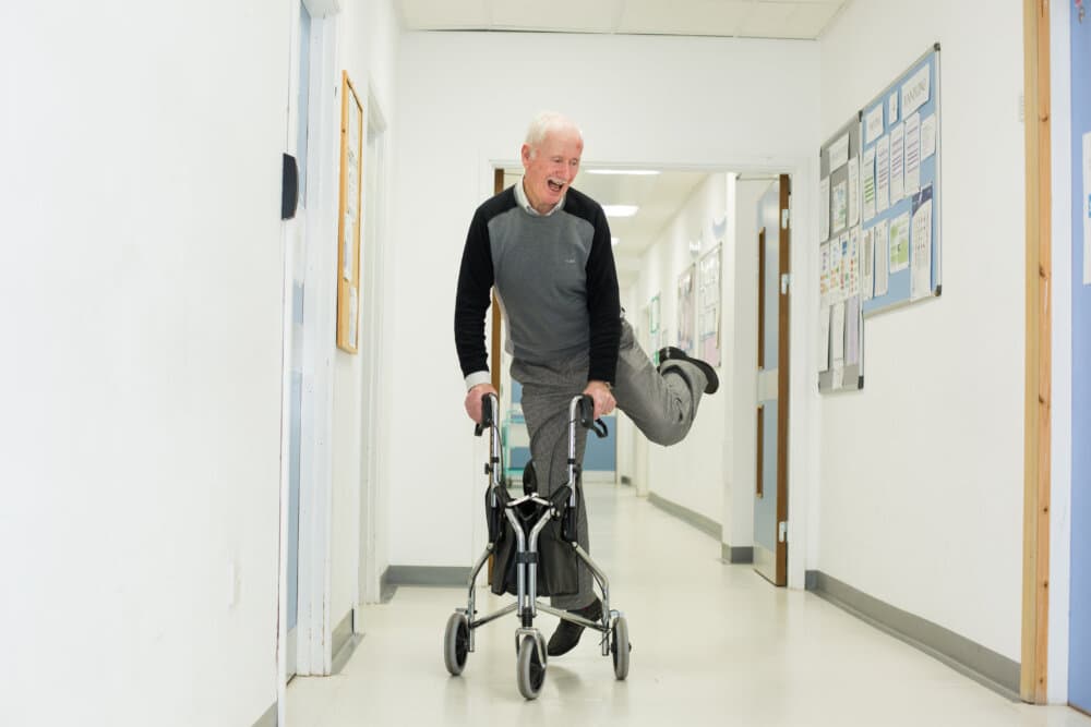 An elderly man playfully balances on one leg using a walker in a bright, cheerful hallway. - Home Instead