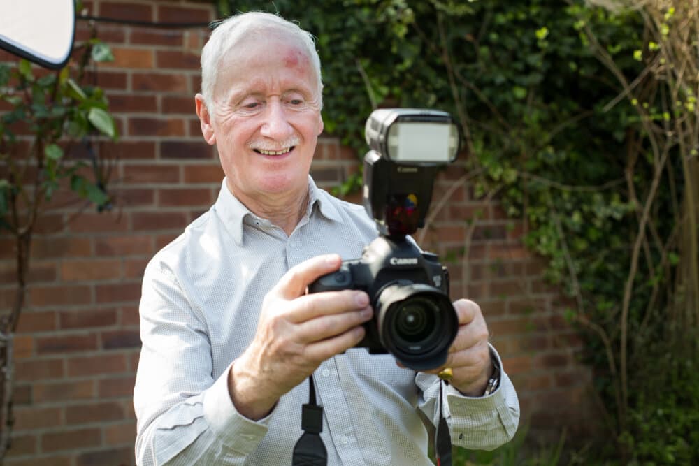 An elderly man in a garden setting smiles while adjusting settings on his camera equipped with a flash. - Home Instead