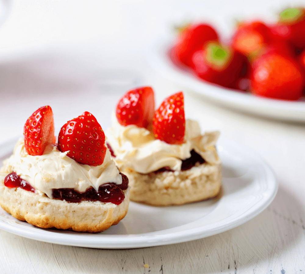 Two scones topped with strawberry jam, clotted cream, and fresh strawberry halves on a white plate, on a light background. - Home Instead