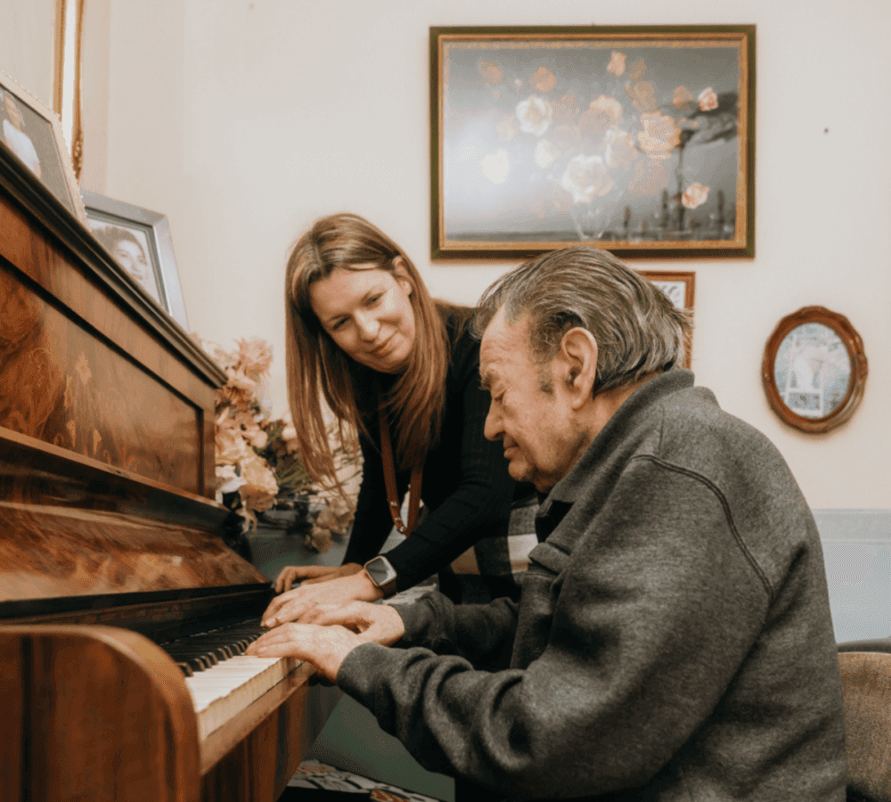 An elderly man plays the piano while a woman leans in to watch in a room with floral pictures on the wall. - Home Instead