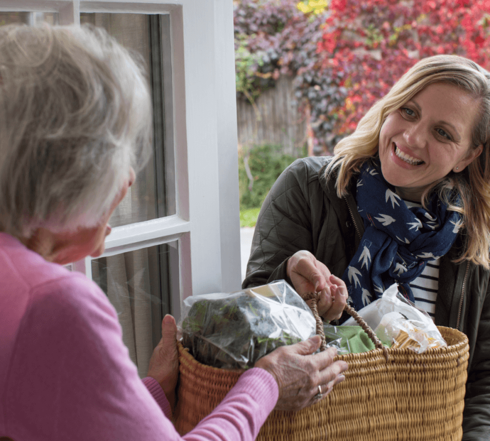 Younger woman hands a basket of groceries to an older woman at the doorway of her home. - Home Instead