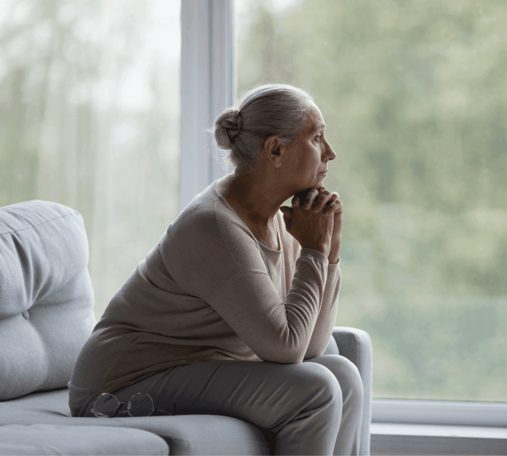 Elderly woman with gray hair sits on a couch, gazing thoughtfully out a window with a green, blurred outdoor background. - Home Instead